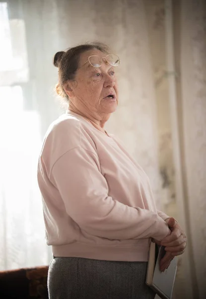 Una anciana mujer caucásica en gafas de pie en el fondo de la ventana con un libro en sus brazos . — Foto de Stock