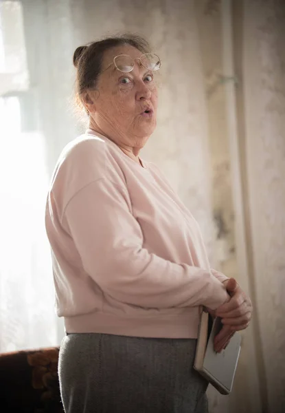 Una anciana con gafas de pie en el fondo de la ventana con un libro en los brazos . — Foto de Stock