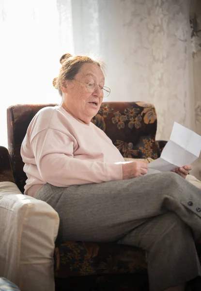 Pasatiempos. Una anciana en gafas disfruta de su pasatiempo sentada en un sillón y leyendo sus cartas en el fondo de la ventana . — Foto de Stock
