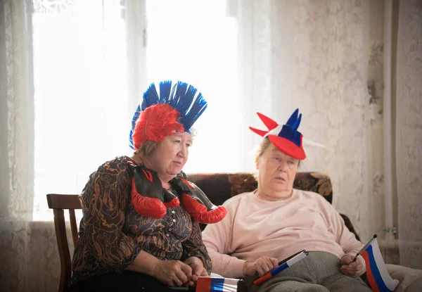 Развлечения. Пожилые женщины поддерживают спортивную команду с флагами, сидящими у телевизора на заднем плане окна — стоковое фото