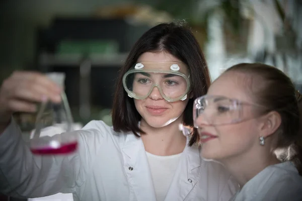 Laboratorio químico. Dos mujeres jóvenes sosteniendo un frasco con líquido rosa en él, sonriendo —  Fotos de Stock