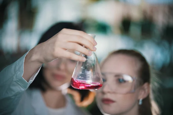 Laboratorio químico. Dos mujeres jóvenes sosteniendo un frasco con líquido rosa en él. Frasco en foco —  Fotos de Stock