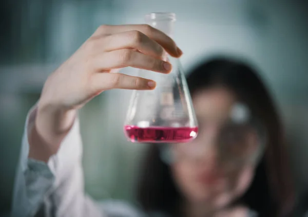 Laboratorio químico. Mujer joven sosteniendo un frasco con líquido en él. Frasco en foco —  Fotos de Stock