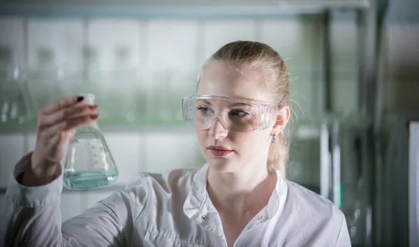 Laboratorio químico. Mujer rubia joven sosteniendo un frasco con líquido en ella. Mujer en foco —  Fotos de Stock