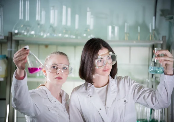 Laboratorio químico. Dos mujeres guapas jóvenes sosteniendo diferentes frascos con líquidos en ella. Mirando el frasco —  Fotos de Stock