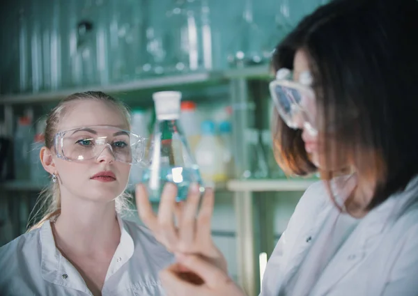 Dos mujeres jóvenes en laboratorio químico sosteniendo un frasco con líquido transparente. Mujer rubia en foco —  Fotos de Stock