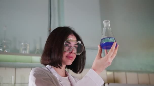 Junge Frau im Chemielabor hält einen Kolben in der Hand und schaut zu — Stockvideo