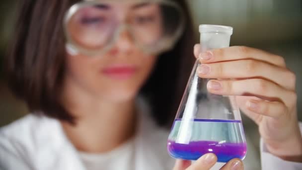化学研究室では青、紫の液体とフラスコを保持している若い女性。フラスコに焦点を当てる. — ストック動画