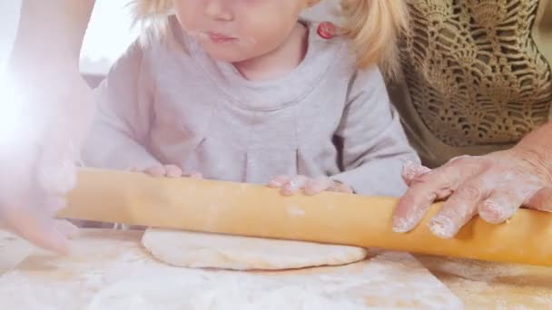 Familia haciendo pastelitos. Una niña pequeña saca la masa con la ayuda de la abuela — Vídeo de stock