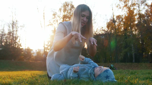 Joven madre feliz y su pequeño bebé jugando en el parque. El bebé acostado en la hierba — Foto de Stock