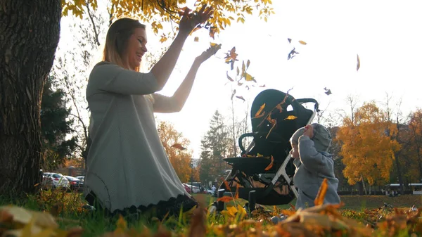 Ung mamma med hennes skrattande lilla bebis kasta löv i luften i höst park — Stockfoto