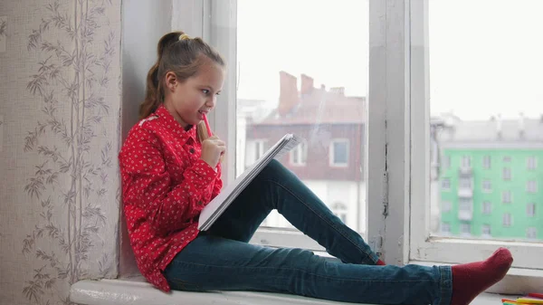 Una bambina seduta sul davanzale della finestra e disegnare, sorridente sul retro della città — Foto Stock