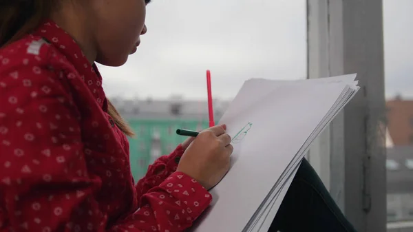 Маленькая девочка сидит на подоконнике и рисует, улыбаясь на заднем плане города. Фокус на бумаге — стоковое фото