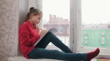 Pencere kenarında oturan ve renkli kalemler ile çizim kırmızı gömlekli bir kız