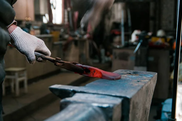 Blacksmith working with red hot metal workpiece of new axe on the anvil at the forge. Focus on hands