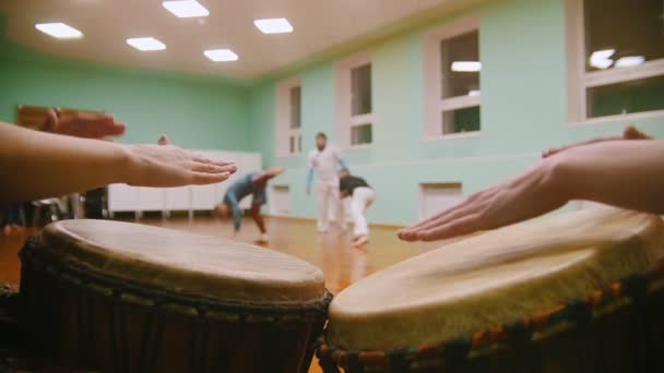 Tocar dos instrumentos musicales jembe o atabaque en el fondo de combate masculino realiza trucos marciales con elementos de baile en el gimnasio deportivo — Vídeos de Stock