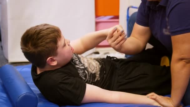 Centro médico. Una doctora ayuda a un niño con parálisis cerebral a levantarse. Hacer ejercicios en la espalda — Vídeos de Stock