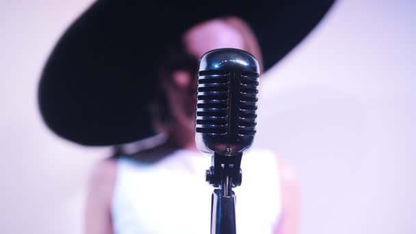 Молодая привлекательная женщина стоит рядом с микрофоном на фоне белой стены в большой шляпе. Мик в фокусе — стоковое видео