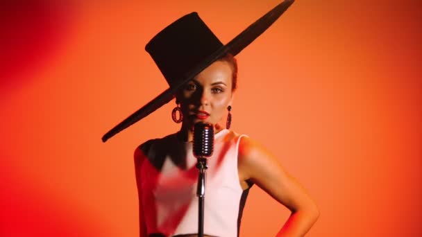 An attractive woman in big retro hat singing on the stage on orange background. — Stock Video
