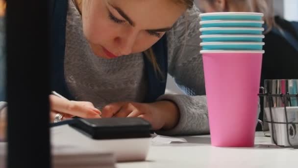 Une femme barista écrit quelque chose sur le journal. Tasses colorées au premier plan . — Video