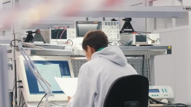 在实验室工作的电子工程师。一位工程师坐在桌旁, 在电脑上工作, 看着报纸. — 图库视频影像