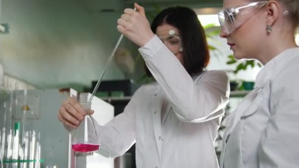 Chemical laboratory. Two young women making experiments. Red liquid in flask — Stock Video