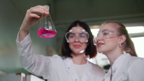 Laboratório químico. Duas jovens mulheres olhando para o frasco com líquido rosa nele — Vídeo de Stock