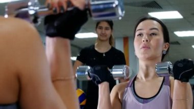 Genç bir kadın el kasları bir dumbbells ile pompalar. Aynaya bakıyorum. Eğitmen tarafından izlenen