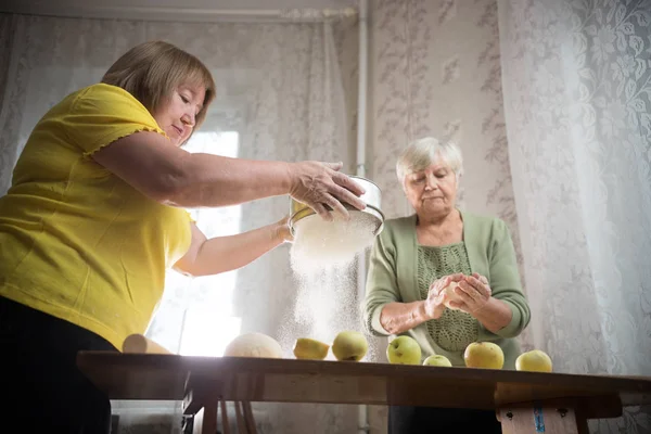 Two old women are making little apple pies. Sifting the flour