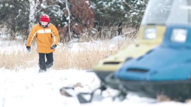 El niño corre a través de las corrientes de nieve en la dirección de una moto de nieve en el bosque — Vídeo de stock