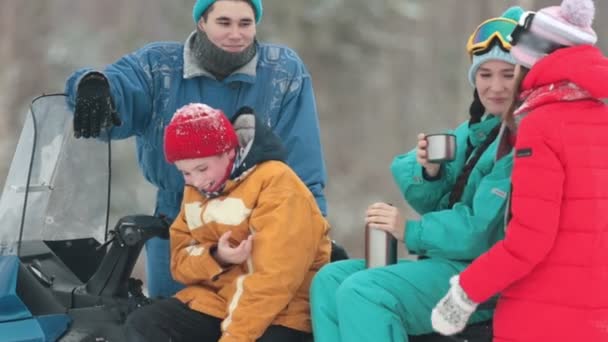 Зима. Семья в разноцветной одежде сидит на снегоходе и пьет горячие напитки из термоса. Маленький мальчик смеется — стоковое видео
