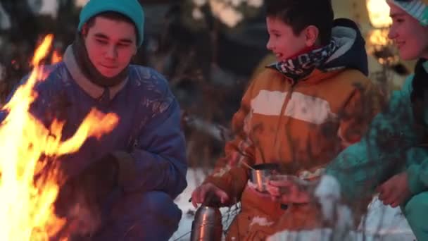 Bosque de invierno. Joven familia feliz sentada en el bosque junto al fuego, bebiendo bebidas calientes del termo y freír salchichas — Vídeo de stock