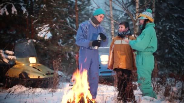 Bosque de invierno. Joven familia parada en el bosque junto al fuego y bebiendo bebidas calientes del termo . — Vídeos de Stock