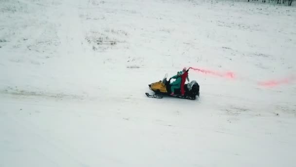 Jóvenes montando motos de nieve en un bosque de invierno. Una persona sosteniendo una bomba de humo roja. Disparos en helicóptero . — Vídeo de stock