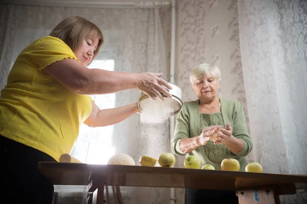 Две пожилые женщины делают маленькие яблочные пирожки. Просеивание муки — стоковое фото