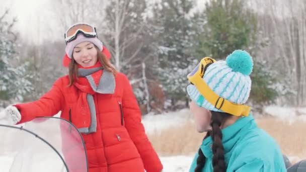 Winter. zwei junge Frauen in bunten Jacken reden und lachen, setzen dann eine Brille auf und bereiten sich auf die Fahrt vor — Stockvideo