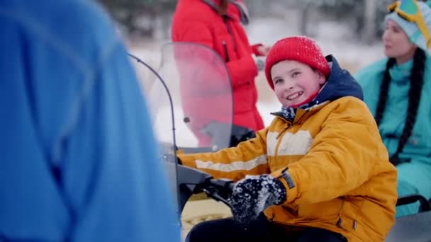 Bosque de invierno. Un niño sonriente sentado en la moto de nieve . — Vídeos de Stock