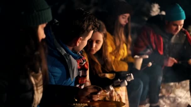 Jovens na floresta de inverno sentados ao lado do fogo. Aquecendo o marshmello perto da fogueira — Vídeo de Stock