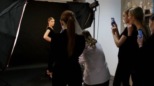 Genç kadın modelinde bir fotoğraf oturumu stüdyoda. Model siyah giysilerle çekim. Tam yüksekliği çekim. Başka modelleri backstage ateş üstünde onların telefon — Stok video