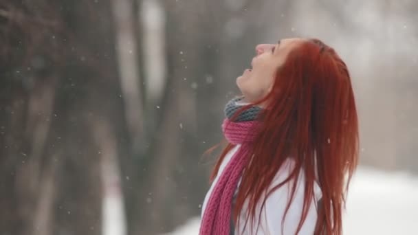 Winter park. Μια γυναίκα με φωτεινά κόκκινα μαλλιά στέκεται στο πεζοδρόμιο. Πιάνει νιφάδες χιονιού με ένα στόμα — Αρχείο Βίντεο