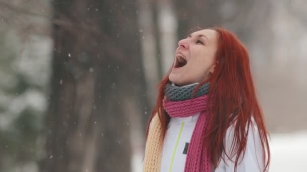 Winter park. Μια γυναίκα με φωτεινά κόκκινα μαλλιά στέκεται στο πεζοδρόμιο. Πιάνει νιφάδες χιονιού με ένα στόμα. Αργή κίνηση — Αρχείο Βίντεο