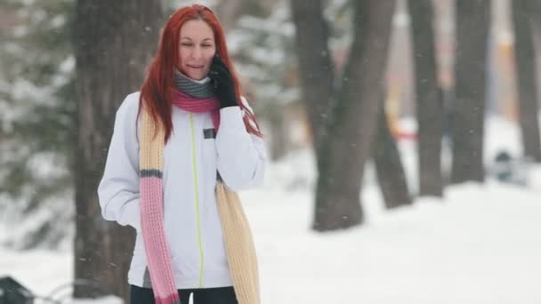 Сильный снегопад. Женщина с яркими рыжими волосами, стоящая в парке и разговаривающая по телефону — стоковое видео