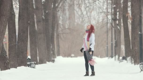 Winter park. A heavy snowfall. A woman with bright red hair standing on the sidewalk. Catching snowflakes with a mouth. — Stock Video