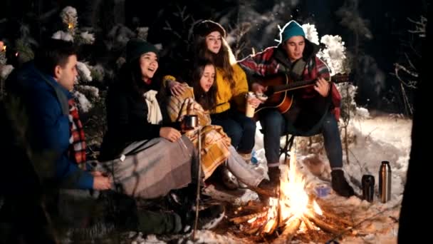 Група щасливих друзів, які сидять в зимовому лісі біля багаття і їдять зефір. Молодий чоловік грає на гітарі. Тепла атмосфера — стокове відео