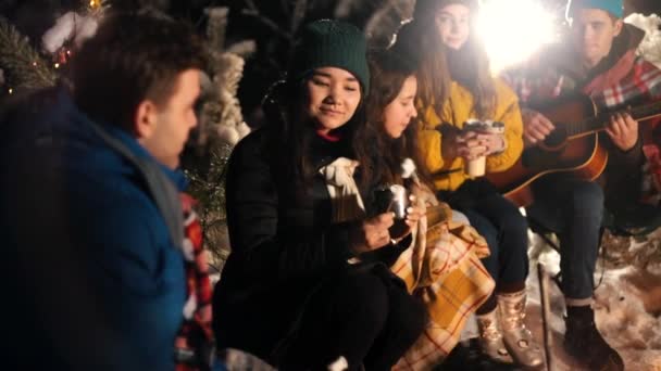 Группа друзей сидит в зимнем лесу у костра и ест зефир. Молодая женщина флиртует и улыбается — стоковое видео