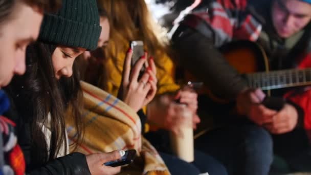 Gruppe von Freunden, die im Winterwald am Feuer sitzen. Sie sitzen in ihren Telefonen. Nahaufnahme — Stockvideo