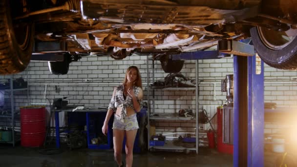 Auto reparatieservice. Een auto-lift. Jonge sexy vrouw in kleine korte broek loopt onder de auto en hangen opknoping op het — Stockvideo