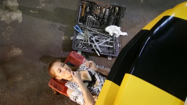 Υπηρεσία επισκευής αυτοκινήτων. Νέοι σέξι γυναίκα βρίσκεται κάτω από το κάτω μέρος του αυτοκινήτου, φωτισμός επάνω το τσιγάρο και έρχεται πίσω στη δουλειά — Αρχείο Βίντεο