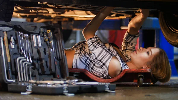 Usługi naprawy samochodów. Seksowny mechanik młoda kobieta leżący pod samochód i praca z klucza. Walizki na narzędzia na pierwszym planie — Zdjęcie stockowe