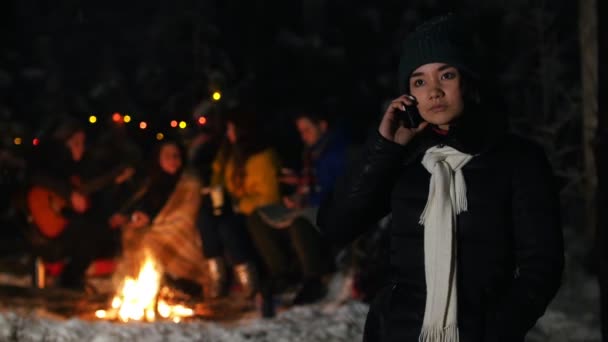 Молода жінка в чорному піджаку стоїть в зимовому лісі і розмовляє по телефону. Група друзів, які сидять біля багаття на фоні — стокове відео